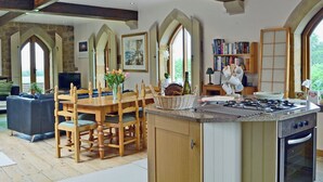 A spacious kitchen with a large island, wooden chairs, and a dining table. There is a stone fireplace, a bookshelf, and a view of the outdoors through large windows.