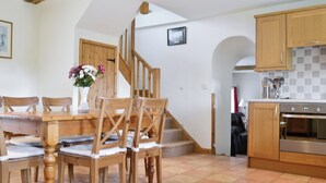 Zona pranzo con sedie e tavolo in legno, cucina con pensili in legno e zoccolo piastrellato, e una scala che conduce a un altro piano.