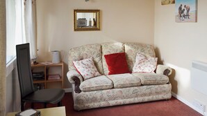 A cozy living room with a floral-patterned sofa, a red pillow, a wooden side table, and a bookshelf. - 2 bedroom accommodation in Dawlish Warren (Dawlish Warren)