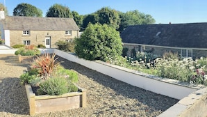 Un bâtiment en pierre doté d’un toit en pente, entouré d’un chemin de gravier et agrémenté d’un jardinier en bois où sont plantées différentes espèces.