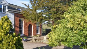Une maison traditionnelle en briques, avec un chemin de pierre menant à l’entrée, entourée d’une végétation luxuriante et d’arbres.
