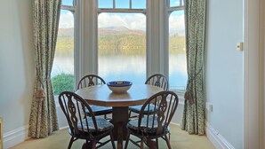 A dining area with a table and chairs, a bowl on the table, and a view of a lake through large windows. - 2 bedroom accommodation in Ambleside (Ambleside)
