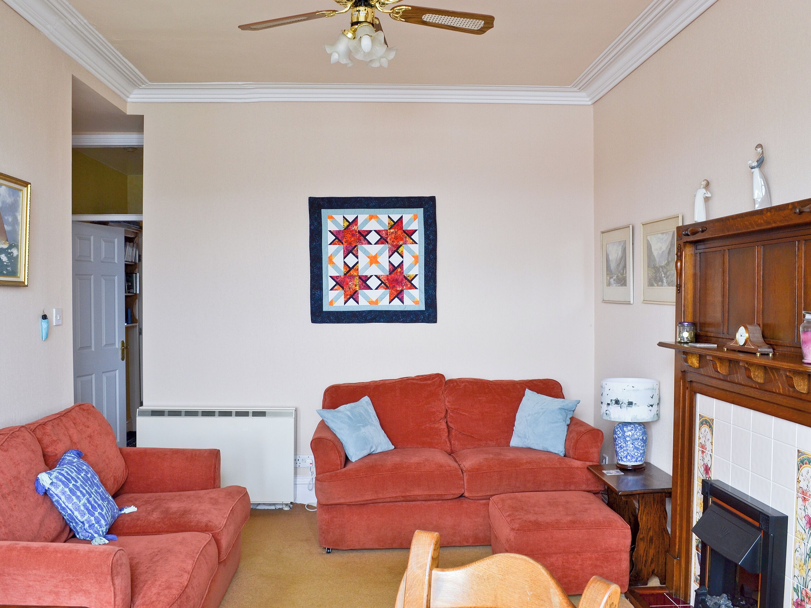 A living room with two red sofas, a ceiling fan, a fireplace, and a quilt on the wall.
