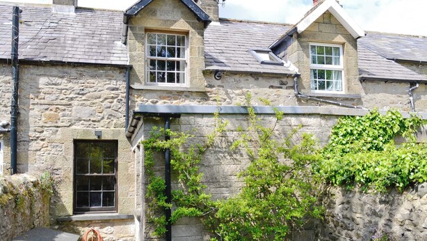 A stone house with two windows, a small porch, and a garden area with a bench. - Langcliffe Lock Cottage (Langcliffe, near Settle)