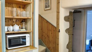 A kitchen with wooden shelves holding kitchenware, a microwave, and a staircase leading to another room. - Langcliffe Lock Cottage (Langcliffe, near Settle)