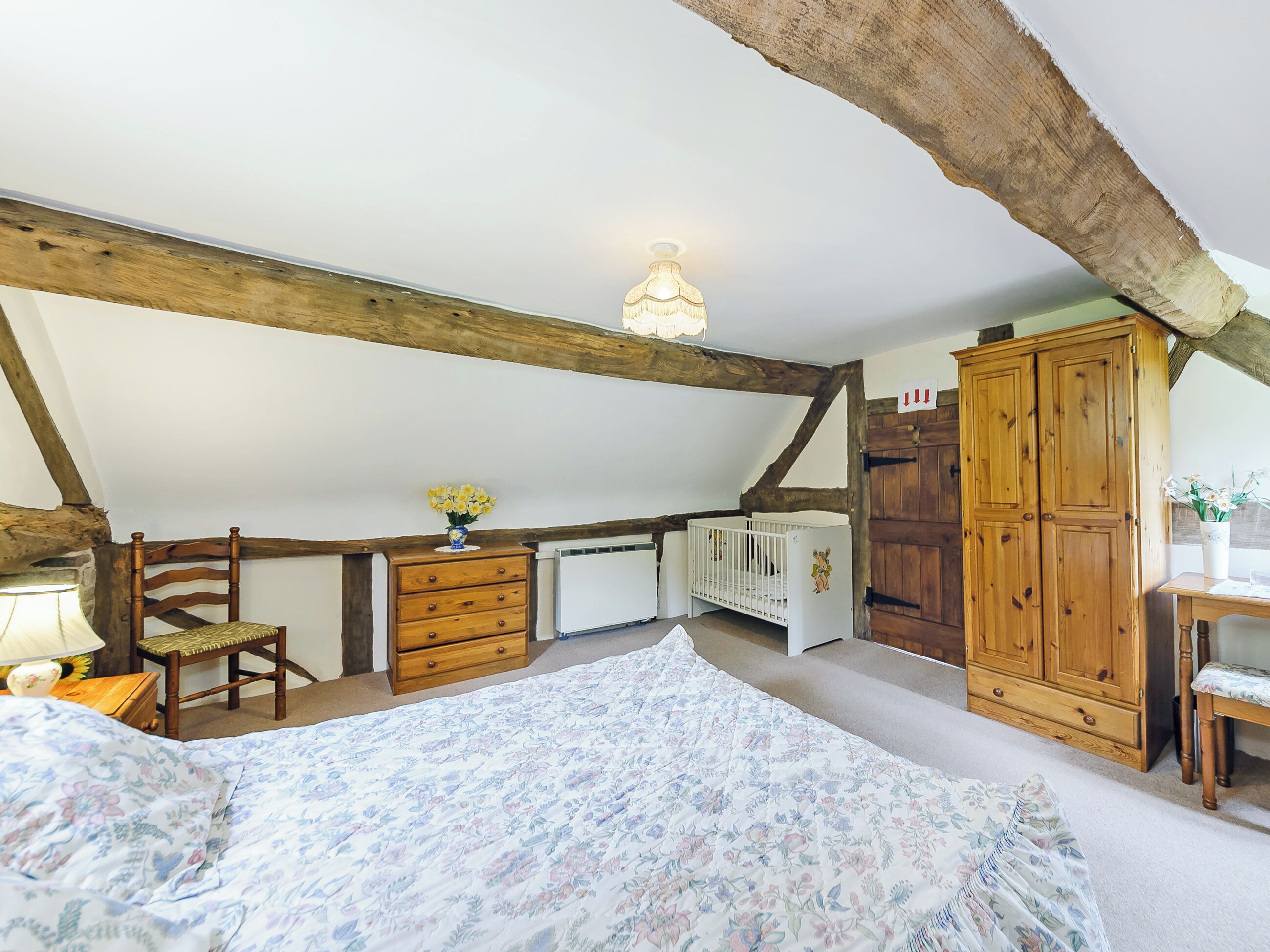 Ein Schlafzimmer mit schrägverdeckelter Decke, Holzbalken, einem Kinderbett, einem Schminktisch, einem Kleiderschrank und einem Nachttisch mit Lampe.