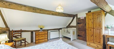 Ein Schlafzimmer mit schrägverdeckelter Decke, Holzbalken, einem Kinderbett, einem Schminktisch, einem Kleiderschrank und einem Nachttisch mit Lampe.