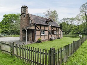 Une maison traditionnelle à colombages, avec une cheminée en pierre, une clôture en bois et une cour couverte d’herbe.