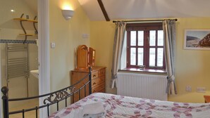 A bedroom with a bed, a dresser, a radiator, a window with curtains, and a bathroom with a towel rack. - Barfield's Cherry Tree Cottage (Canworthy Water, near Crackington Haven)