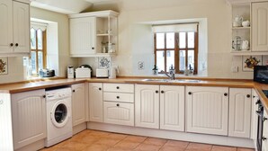 A kitchen with white cabinetry, a washing machine, and a window with curtains. - Barfield's Cherry Tree Cottage (Canworthy Water, near Crackington Haven)