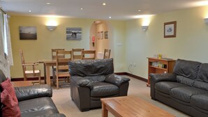 A living room with leather sofas, a wooden coffee table, and a dining area with wooden chairs and a table. - Barfield's Cherry Tree Cottage (Canworthy Water, near Crackington Haven)