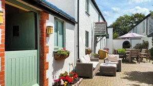 A courtyard with outdoor seating, a brick wall, and hanging flower baskets. - Barfield's Cherry Tree Cottage (Canworthy Water, near Crackington Haven)