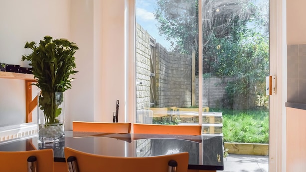 A dining area with a glass table and chairs, a vase with greenery, and a view of a garden through a sliding glass door. - The House On The Green (Chipping Norton)