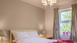 A bedroom with a bed, two bedside tables with lamps, a window with curtains, and a chandelier. - The House On The Green (Chipping Norton)