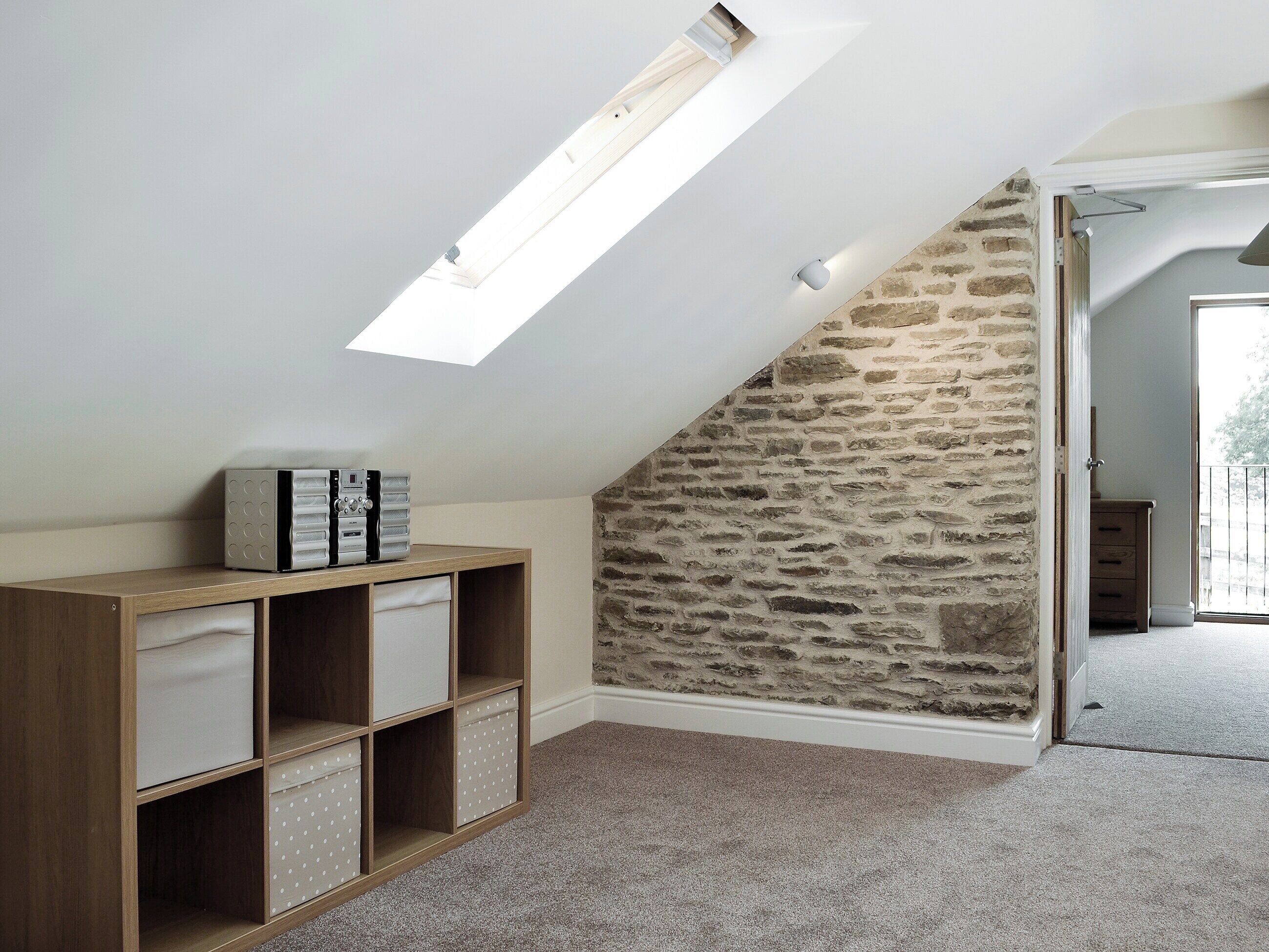 Ein Zimmer mit geneigter Decke, einem Oberlicht, einem Holzregal und einer Steinwand.