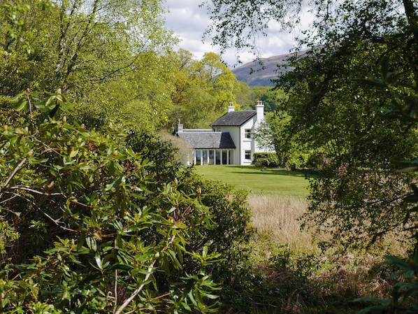 A house with a large lawn, surrounded by dense trees and hills in the background.
