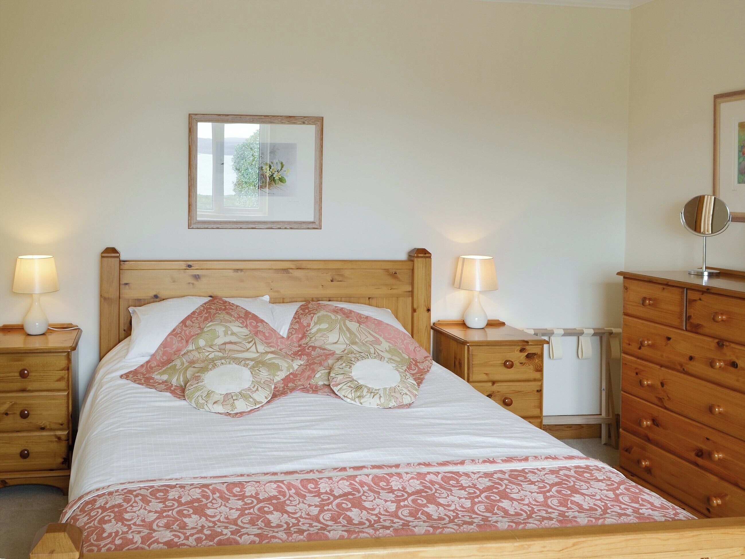 A neatly made bed with floral bedding, two bedside tables with lamps, a wooden dresser, and a framed picture on the wall.