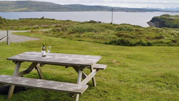Una mesa de picnic con una botella y un vaso encima, con vistas a un lago y colinas al fondo.