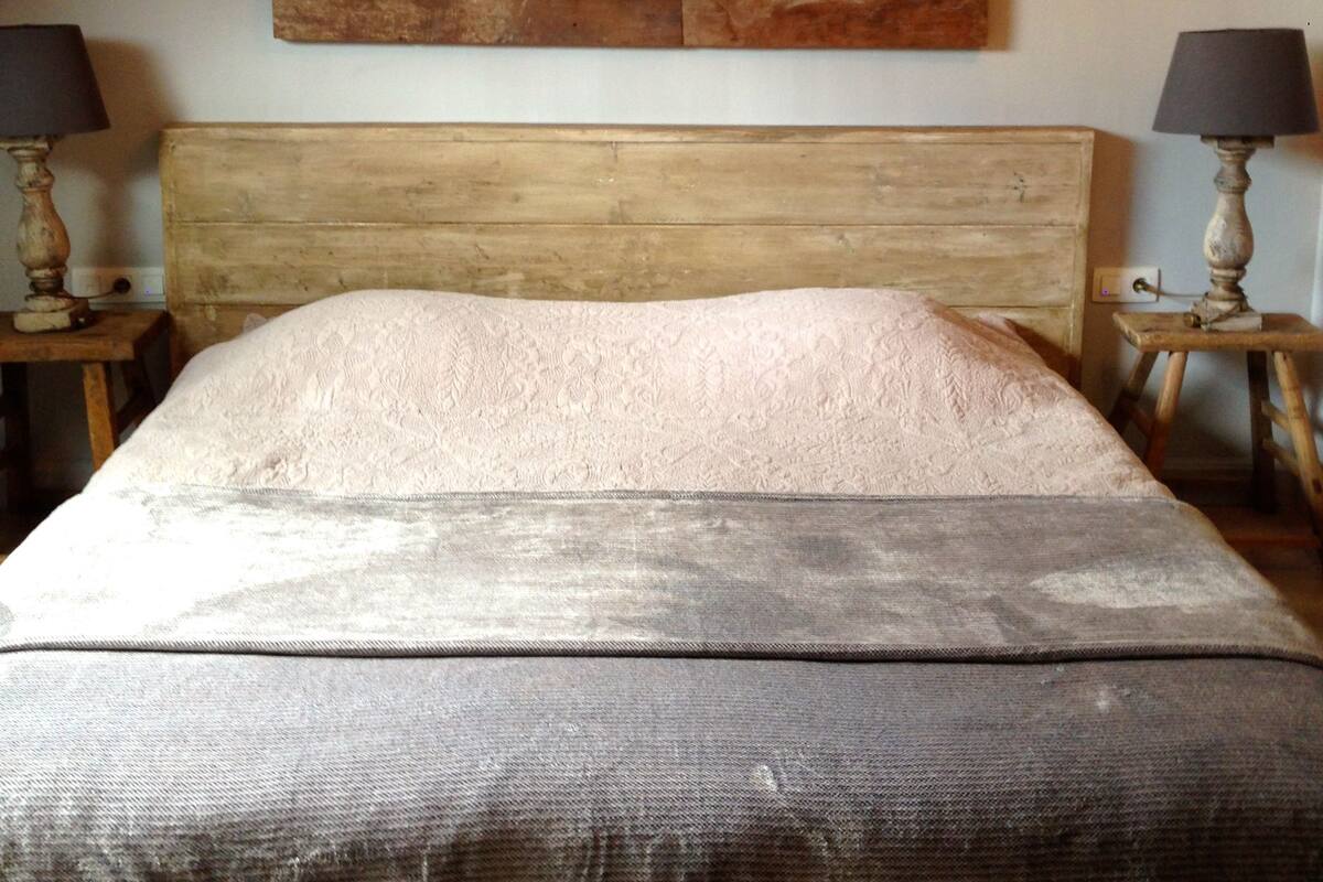 A bedroom with a wooden headboard, a large bed with a textured blanket, two bedside tables with lamps, and a rustic wall art piece.