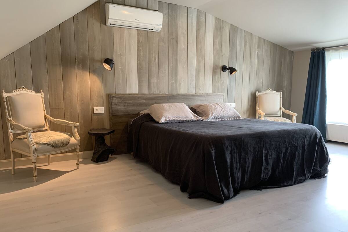 A modern bedroom with a large bed, two chairs, a small table, and an air conditioning unit on the wall.