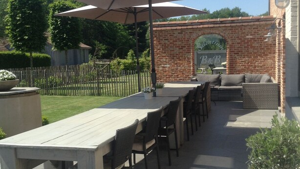 An outdoor patio with a long table and chairs, a large umbrella, and a brick wall with a sign. - B&B Silentium (Tielt-Winge)