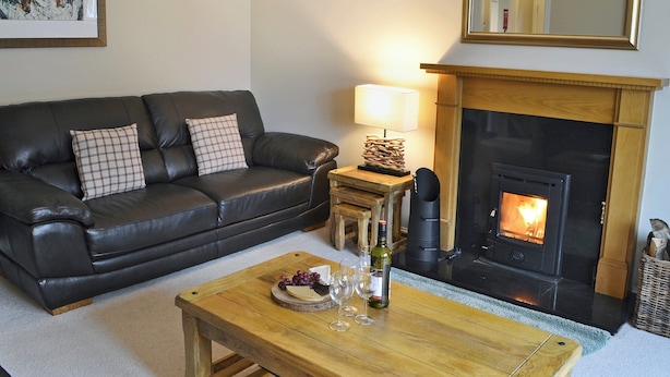 A living room with a black leather sofa, a wooden coffee table with a bottle and glasses, and a fireplace. - 4 bedroom accommodation in Aviemore (Aviemore)
