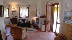 Un salon comprenant un canapé, deux fauteuils en osier, un téléviseur et un coin cuisine visible à travers une porte ouverte.