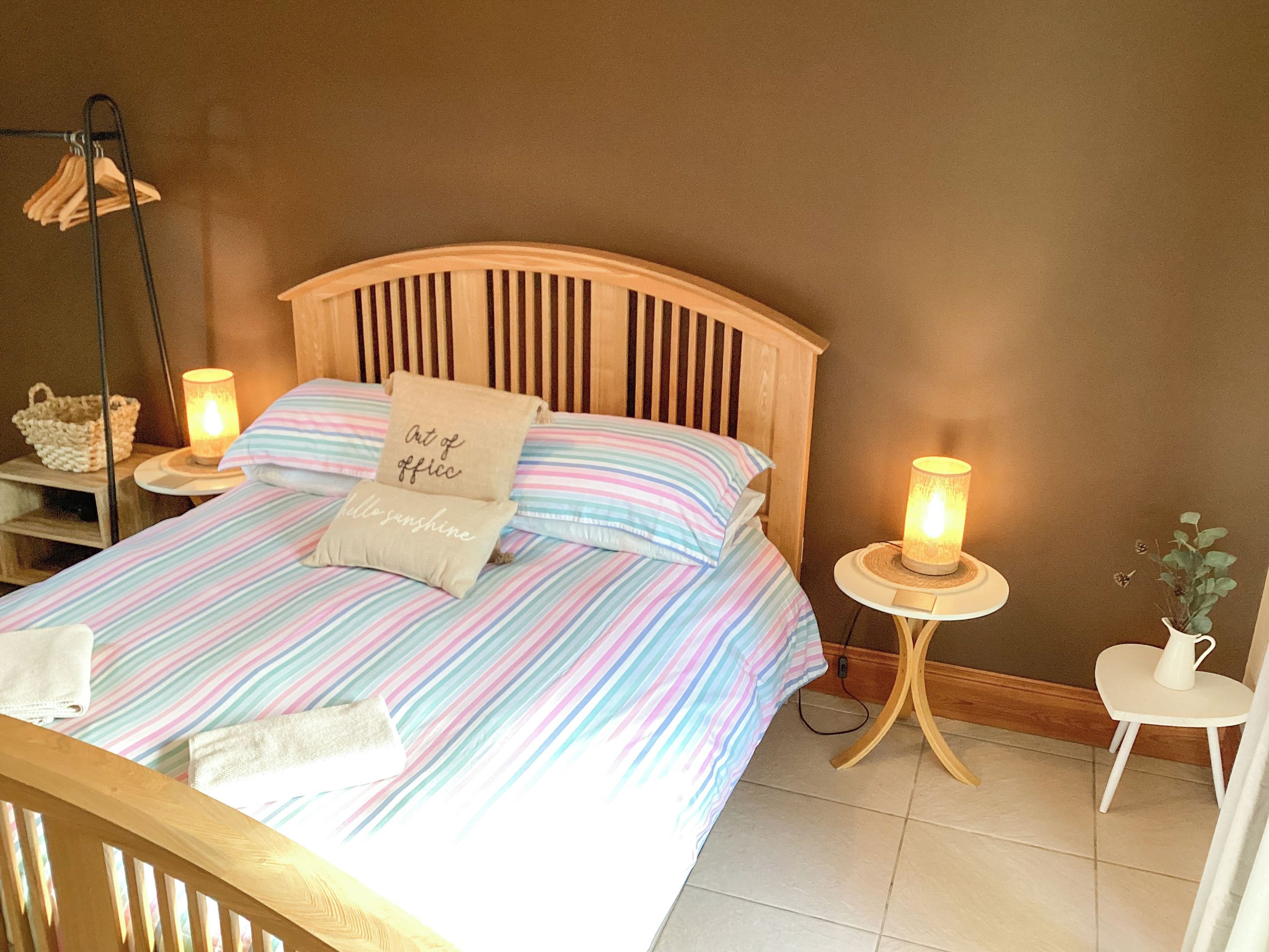 Una cama bien tendida con ropa de cama a rayas, dos lámparas de noche, un cabecero de madera, una mesita pequeña con una lámpara y una cesta de mimbre en una mesita de noche.