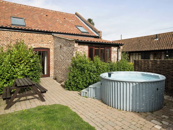 A brick house with a tiled roof, a hot tub, a wooden picnic table, and a grassy area.