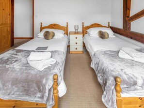 A hotel room with two wooden beds, each with a white mattress and a grey blanket. There is a wooden nightstand with a lamp between the beds.
