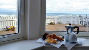 Ett bord med croissanter och en kaffekanna står vid ett fönster med utsikt över stranden.