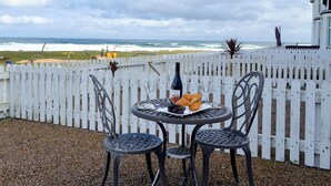 Ett bord med en vinflaska, ett vinglas och mat, placerat framför ett vitt staket med utsikt över havet.