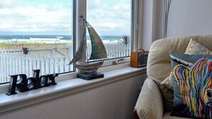 Ett rum med utsikt över havet genom ett fönster, en liten modell av en segelbåt på fönsterbrädan och en bekväm soffa med färgglada kuddar.