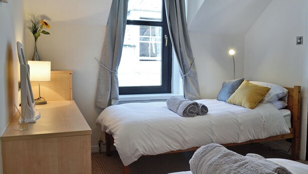 A bedroom with two beds, a bedside table with a lamp, a window with curtains, and a wall-mounted shelf with a vase. - 2 bedroom accommodation in Moffat (Moffat)