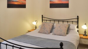 Un lit bien fait, recouvert d’une couverture grise, avec deux oreillers assortis et une tête de lit en métal noir. Deux lampes de chevet sont posées sur des tables de chevet en bois, et deux tableaux représentant des couchers de soleil ornent le mur.