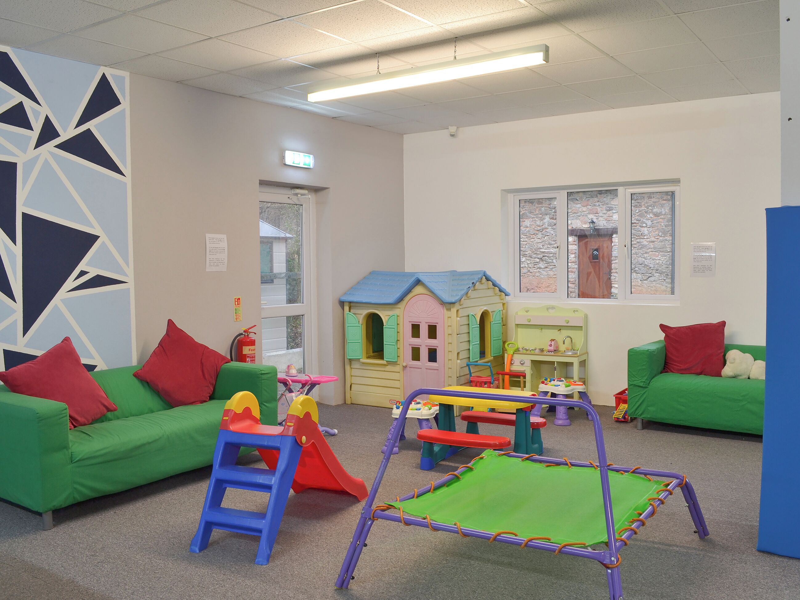 Une salle de jeux avec des canapés verts, une maison de jeu colorée et un petit trampoline.