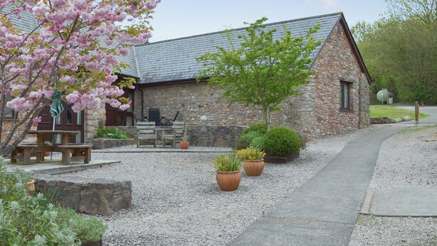 Un bâtiment en pierre avec un toit en pente, une terrasse en bois avec des chaises et des plantes en pot devant.