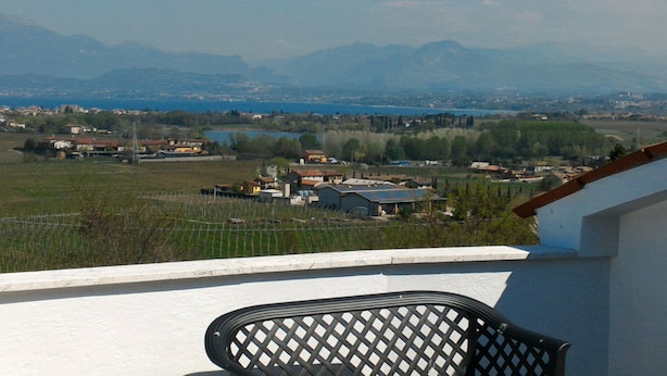 Un balcón con una silla de metal, con vistas a un paisaje rural que incluye casas, un lago y montañas a lo lejos.