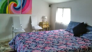 Un dormitorio con una colcha estampada de flores, una silla blanca, una mesita de noche con una lámpara y una ventana con cortinas.