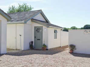 A small, single-story building with a grey door and a small window. There are potted plants on the ground and a gravel driveway.
