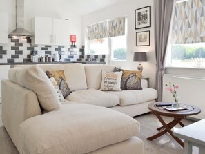 A modern living room with a beige sectional sofa, a round wooden coffee table, and a kitchen in the background.