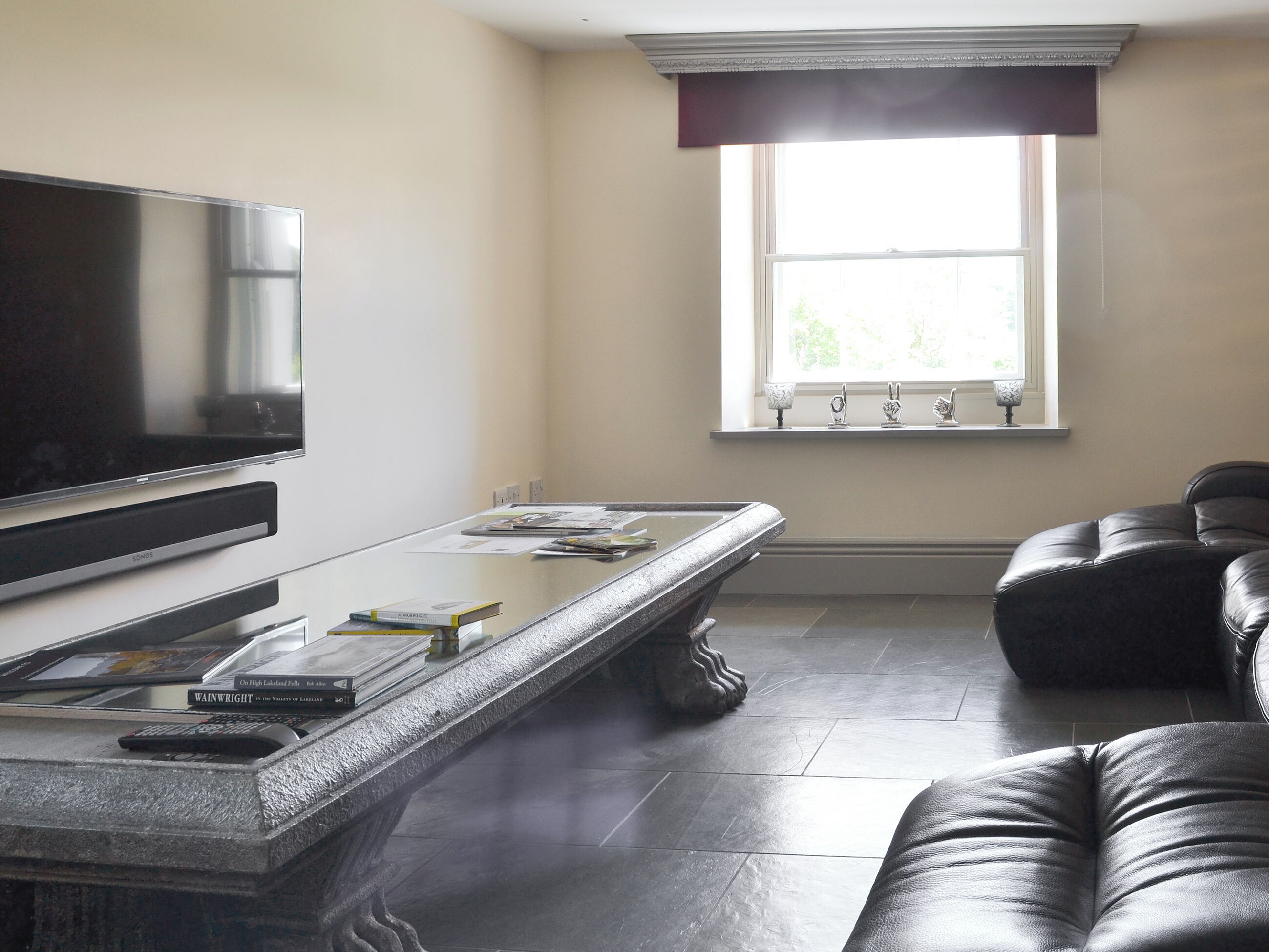 A living room with a flat-screen TV, a glass-top coffee table, a leather sofa, and a window with decorative items.
