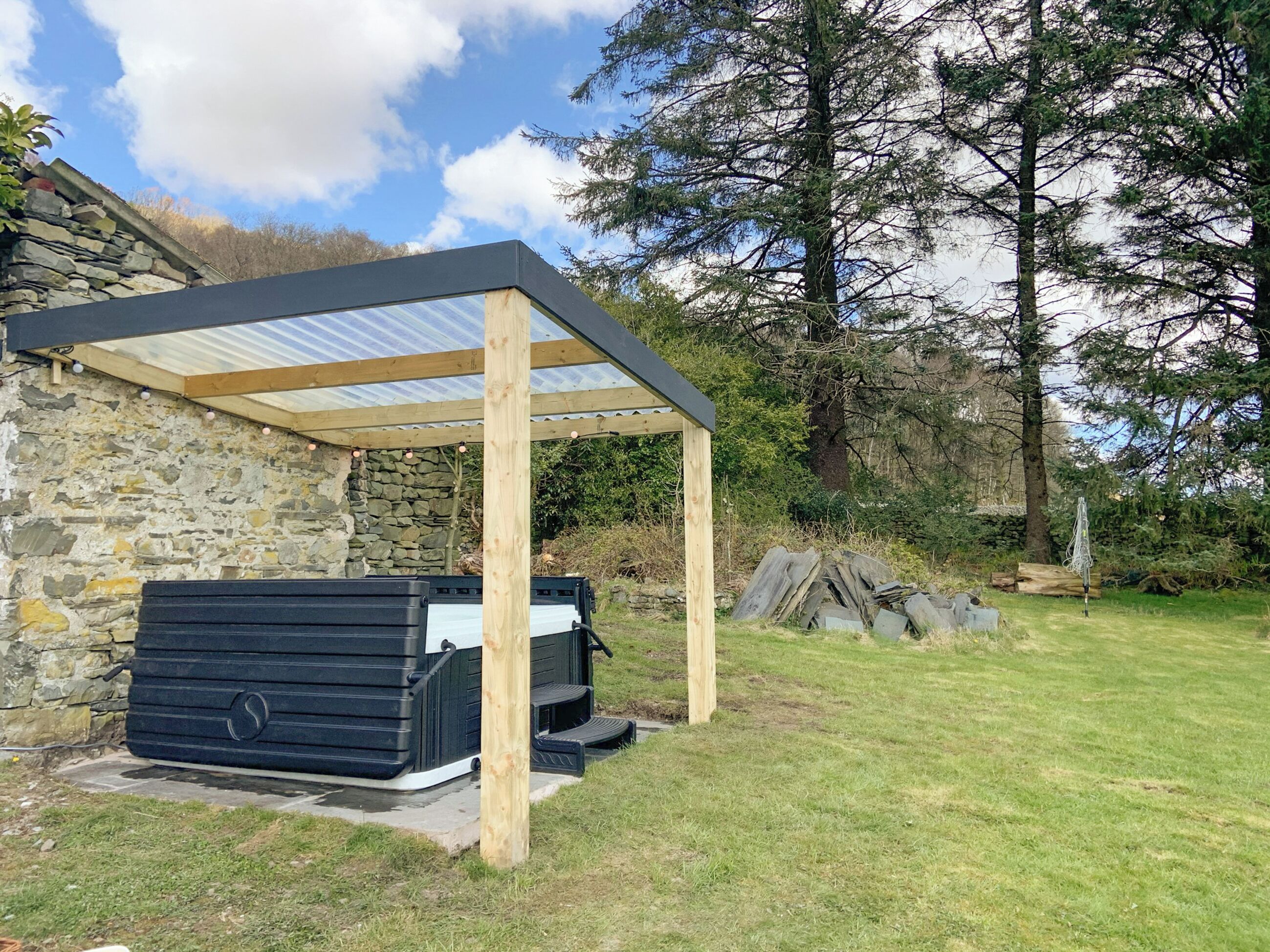 Una banyera d'hidromassatge sota una pérgola de fusta amb parets de pedra, envoltada d'un jardí amb gespa i arbres.