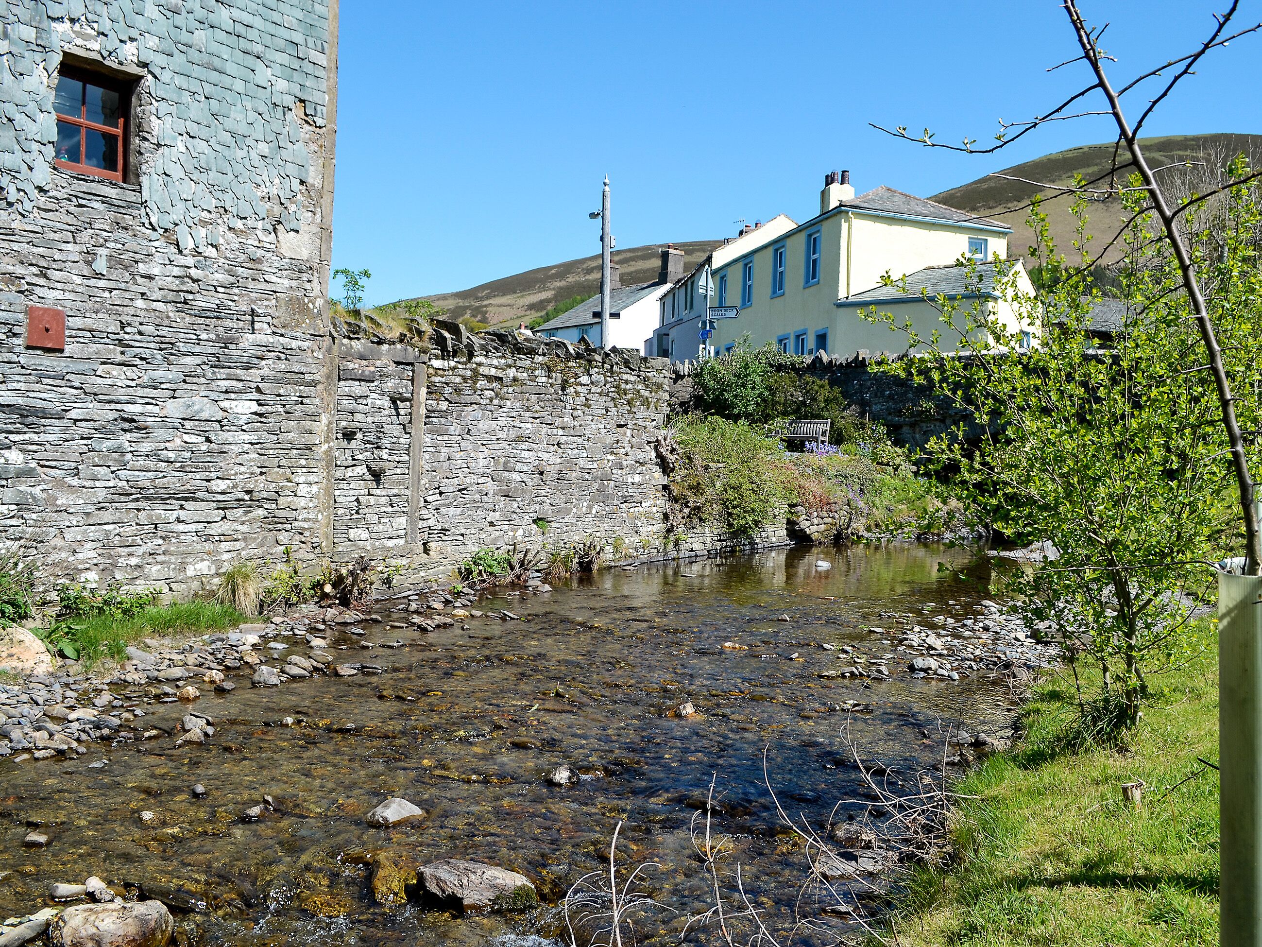 A stone building with a window, a small stream, and a hill in the background.