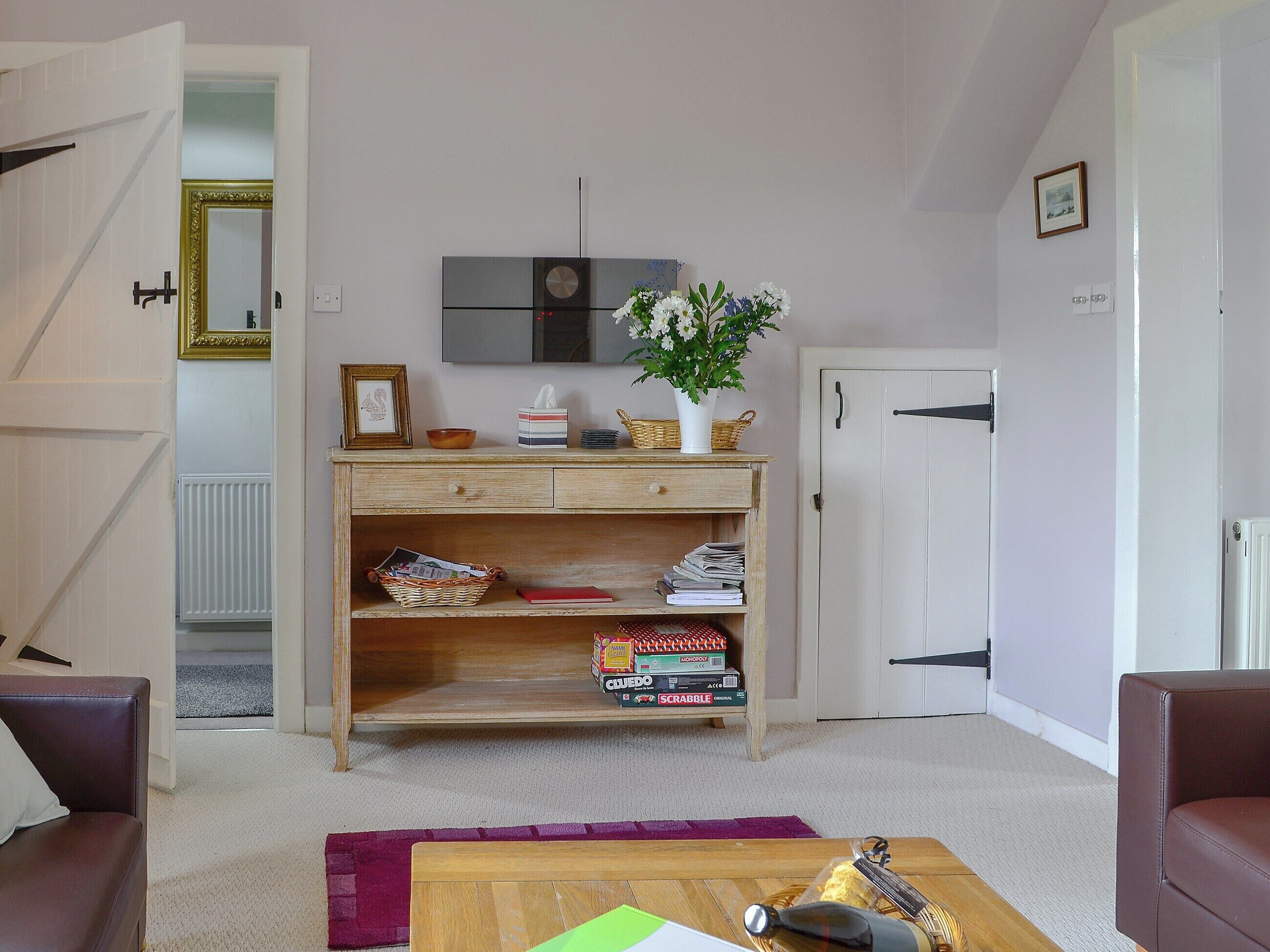 A living room with a wooden coffee table, a red book, and a vase with flowers. There is a bookshelf with board games and a framed picture.