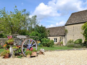 A stone building with a traditional stone roof, surrounded by a gravel driveway and a garden with potted plants and a wooden cart. - 2 bedroom accommodation in Tarlton, Cirencester (Tarlton, Cirencester)