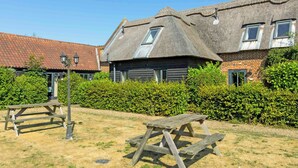 Een traditioneel gebouw met rieten dak en een gedeelte van baksteen, met een houten bankje en een haag op de voorgrond.