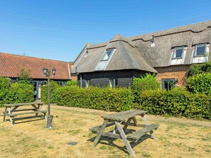 A traditional thatched-roof building with a brick section, a wooden bench, and a hedge in the foreground.