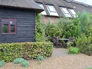 Une maison traditionnelle avec un toit de chaume, un mur en briques et un jardin avec un chemin de gravier menant à une table et des chaises en bois.