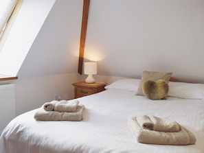 Un dormitorio con techo inclinado, una mesita de noche con lámpara, cama con ropa blanca y dos toallas enrolladas sobre la cama.